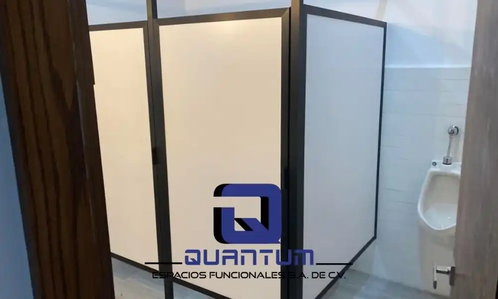 Mamparas sanitarias en blanco y negro para un estilo moderno Vista de un sanitario con mingitorio blanco y mamparas sanitarias de color blanco con perfiles de aluminio en color negro. El diseño moderno y contrastante crea un ambiente limpio y actual. El logo de "Quantum Espacios Funcionales S.A. de C.V." se encuentra en la parte inferior central de la imagen.