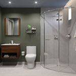 Diseño de baño moderno con pared verde olivo, cancel para ducha de vidrio templado curvo y sanitario blanco. Servicio de instalación de canceles en Tlaquepaque, Jalisco.